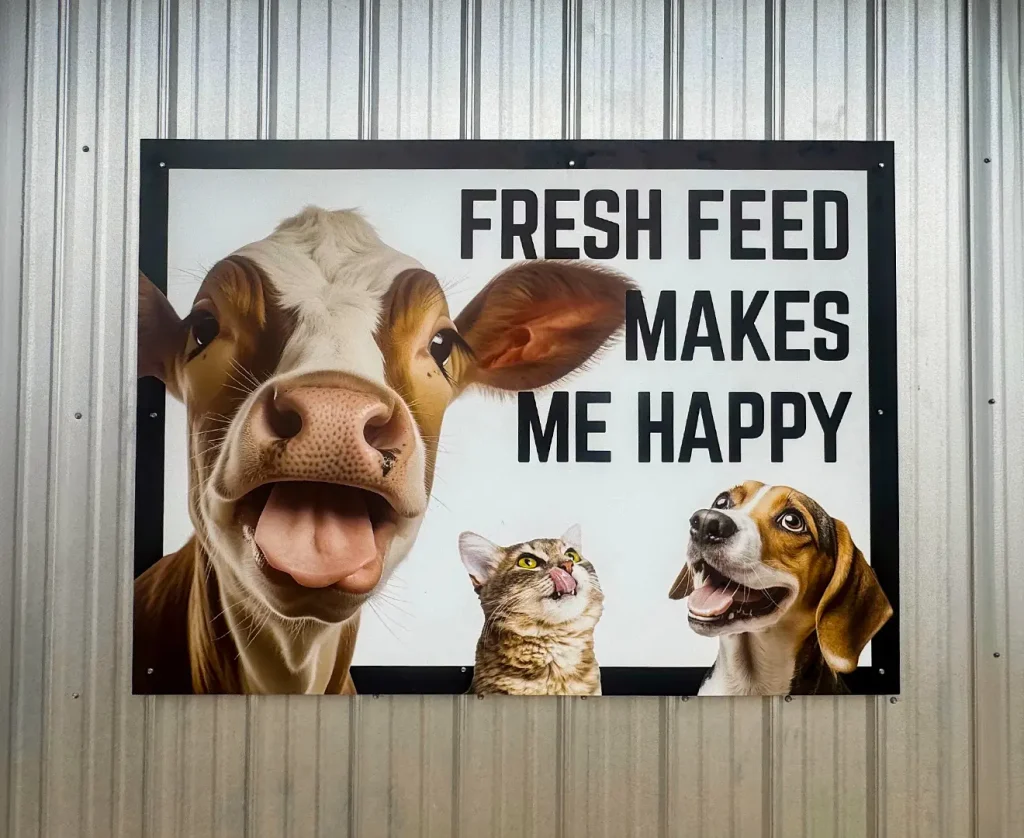 Exterior sign at Valley Feed & Supply with cow, cat, and dog illustration reading “Fresh Feed Makes Me Happy.”
